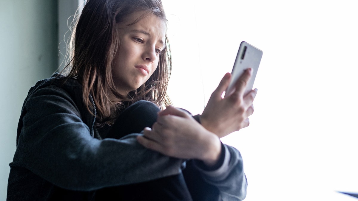 Teenage girl sitting at the window and looking at her phone with a sad expression