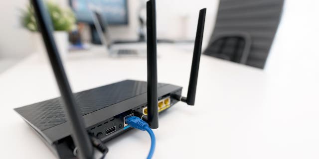 Bilgisayar monitörlerinin yanındaki beyaz masada siyah Wi-Fi yönlendirici