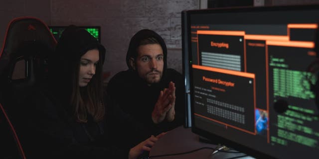 A girl and a guy in black hoodies sit near computer screens hacking 