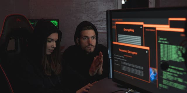 Girl and guy in black hoodies sitting next to computer screens hacking 