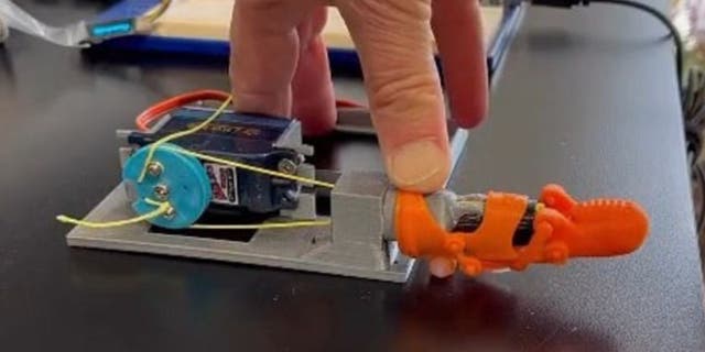 Orange 3D-printed prosthetic pinky on a contraption with man's hand on top