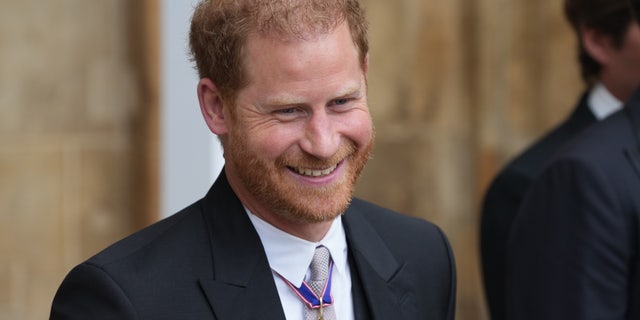 Prince Harry smiles in a black suit and white button down with medals adorned on his suit at the coronation of King Charles