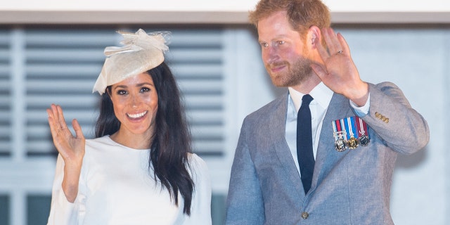 Meghan Markle wearing a creme dress and a matching hat next to Prince Harry in a grey suit and black skinny tie