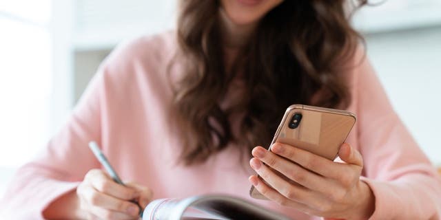 Woman in pink shirt smiling and holding phone while writing in journal with a pen