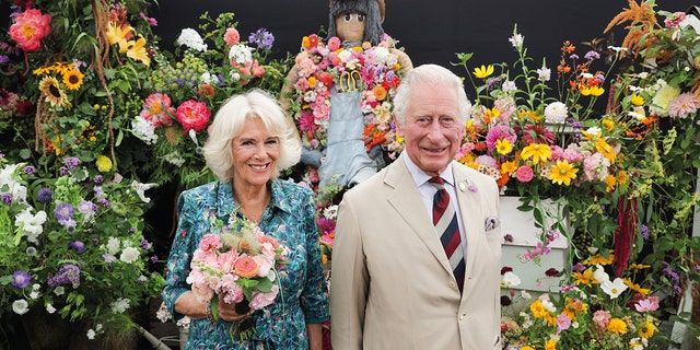 Camilla in a printed blue dress next to King Charles in a beige suit surrounded by flowers
