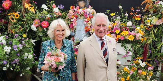 Camilla in a printed blue dress next to King Charles in a beige suit surrounded by flowers