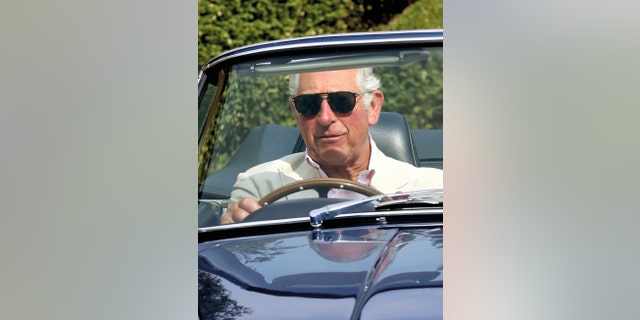 King Charles in a beige suit and sunglasses driving his vintage car