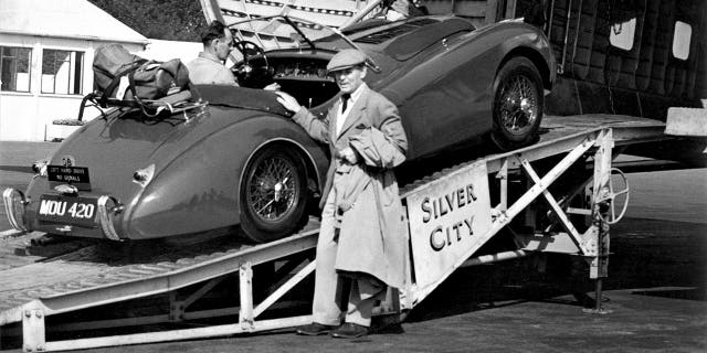 Clark Gable in a suit hold onto a Jaguar car as it is loaded aboard a plane