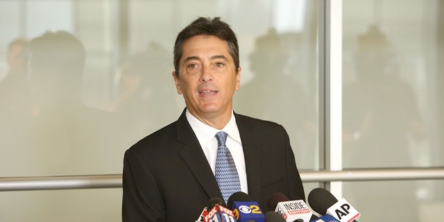 Scott Baio behind podium in a black suit, white button-down and blue tie