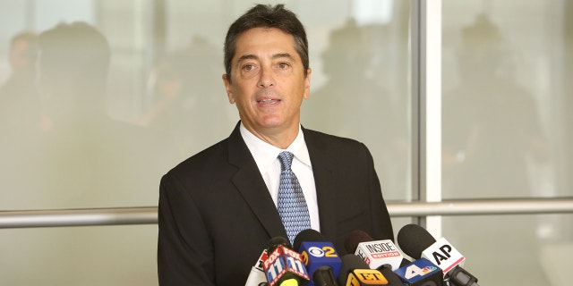 Scott Baio behind podium in a black suit, white button-down and blue tie