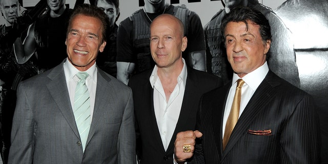 Arnold Schwarzenegger in a grey suit and green tie stands next to Bruce Willis with a white shirt and black jacket next to Sylvester Stallone flashing a fist in a black suit and gold tie at the premiere of "The Expendables"