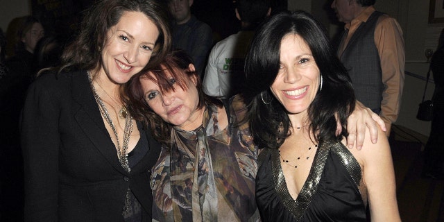 Joely Fisher in black and a long chain necklace is embraced by Carrie Fisher in a patterned top leaning her head on Joely's shoulder, next to Tricia Leigh Fisher also in a black v-cut top