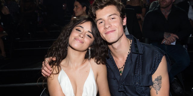 Camila Cabello in a white dress smiles as Shawn Mendes wraps hi arm around her