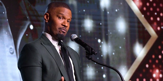 Jamie Foxx looks at the camera behind the podium at the AAFCA Awards wearing a dark suit and black tie