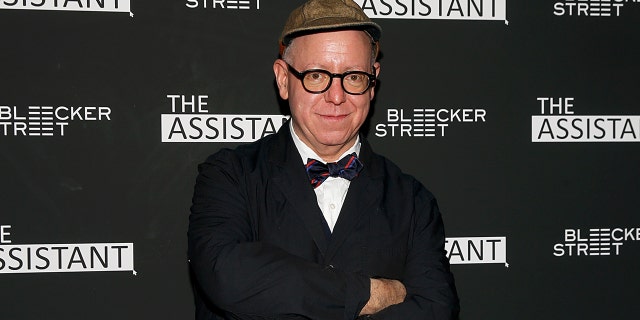 Writer James Schamus soft smiles for the camera at a screening of "The Assistant" wearing a suit and bow-tie, with his arms crossed, dark rimmed glasses and a hat