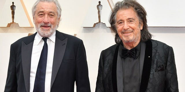 Robert De Niro smiles in a black suit and tie next to Al Pacino in a black suit at the Academy Awards in 2020