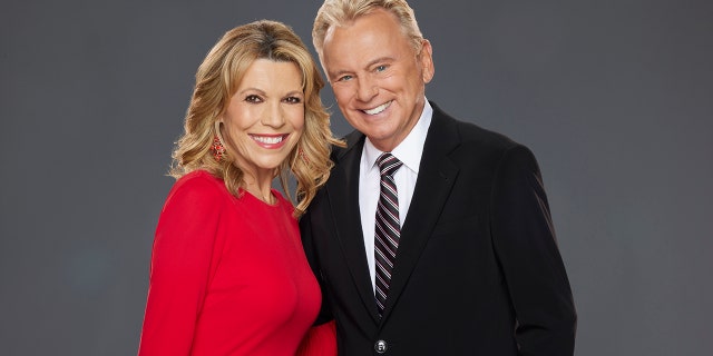 Vanna White in a red dress smiles in a promotional photo for "Celebrity Wheel of Fortune" with Pat Sajak in a black suit and striped tie