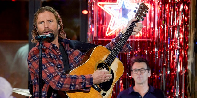 Dierks Bentley in a red plaid holds up a guitar and strums in front of a microphone