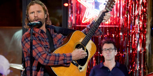 Dierks Bentley in a red plaid holds up a guitar and strums in front of a microphone