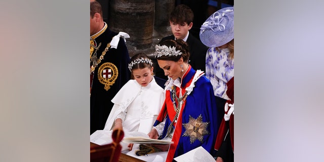 Inside Westminster Abbey, Princess Charlotte in white and a leaf tiara looks at a reading with her mother Catherine, Princess of Wales, in a blue robe with red ribbon and a matching tiara