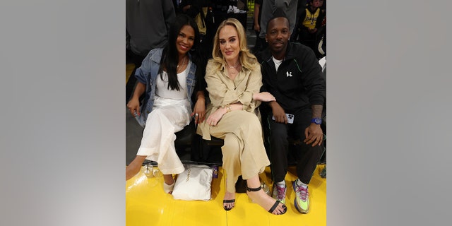 Nia Long in white and a denim jacket poses next to Adele in a beige jumpsuit, holding the arm of her boyfriend Rich Paul, in black, seated courtside
