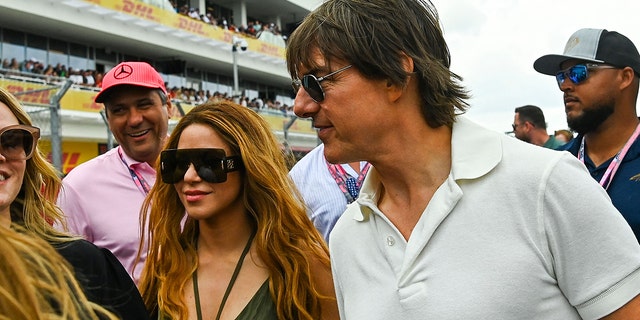 Shakira in a black tank top with two strings in the center around her neck and massive glasses walks alongside Tom Cruise in a white polo shirt at the Grand Prix in Miami