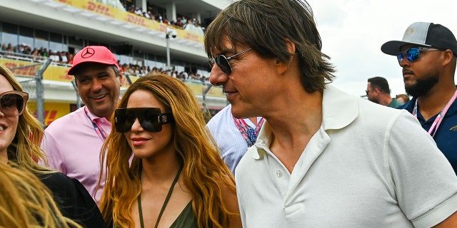 Shakira in a black tank top with two strings in the center around her neck and massive glasses walks alongside Tom Cruise in a white polo shirt at the Grand Prix in Miami