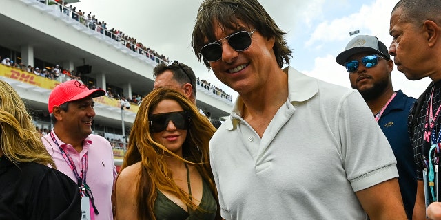 Shakira in a black tank top with two strings in the center around her neck and massive glasses soft smiles for a picture with Tom Cruise in a white polo shirt at the Grand Prix