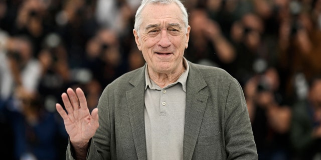 Robert De Niro in a light grey shirt and darker grey jacket waves to the crowd in Cannes