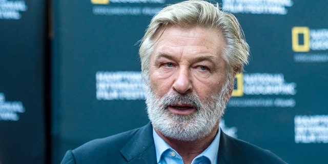 Alec Baldwin with a beard looks off camera on the red carpet, wearing a blue shirt and blazer