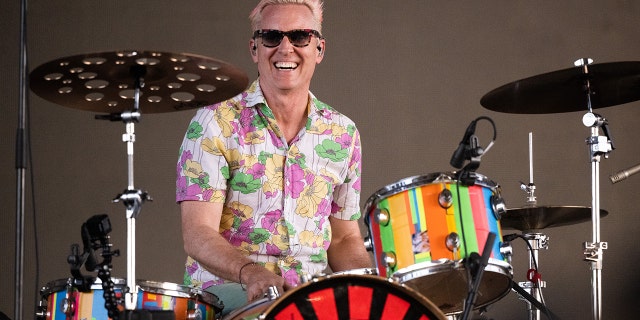 Josh Freese in a colored patterned shirt smiles with sunglasses on behind a set of drums playing at Coachella