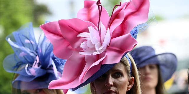 Two women wear large flower-shaped hats at the Kentucky Derby.  One woman wears a pink hat while another woman wears a purple hat.