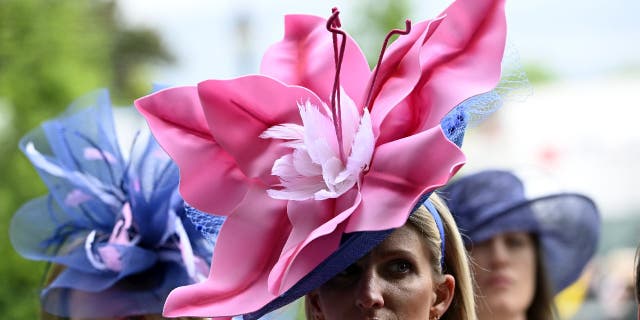 Two women wear large flower-shaped hats at the Kentucky Derby.  One woman wears a pink hat while another woman wears a purple hat.