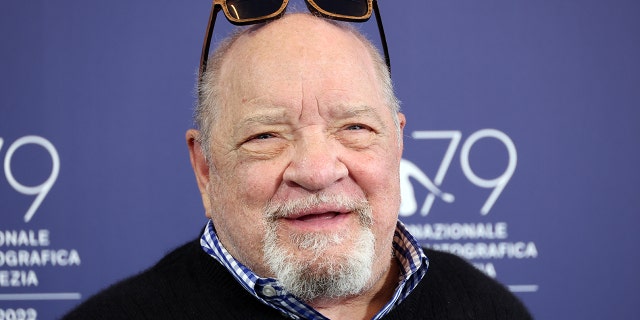 Paul Schrader smiles with his sunglasses on top of his head, wearing a checkered shirt and sweater over on the red carpet