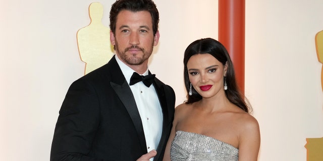 Miles Teller in a tuxedo smiles on the carpet with wife Keleigh in a silver gown at the Oscars