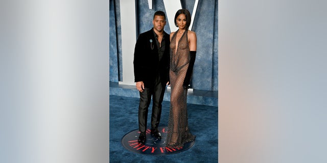 Russell Wilson in a black outfit at the Vanity Fair Oscar Party holds onto Ciara in a mesh/fishnet dress