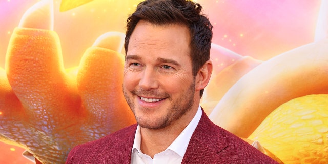 Chris Pratt wearing a white button down and red jacket smiles on the red carpet