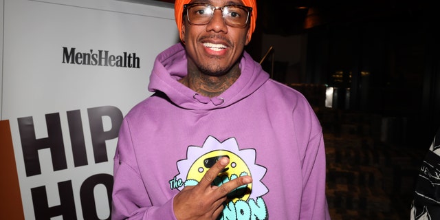 Nick Cannon in a purple sweatshirt throwing up a piece sign and wearing a red cap