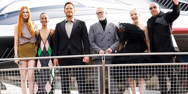 Karen Gillan, Zoe Saldana, Chris Pratt, James Gunn, Pom Klementieff and Vin Diesel stand behind a fence and look out to the crowd at a European gala event at the Avengers Campus in Disneyland Paris while promoting "Guardians of the Galaxy Vol. 3"