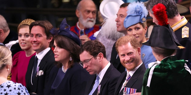 Prince Harry, seated, smiles in a suit as his aunt Anne, Princess Royal approaches him wearing a green jacket and dark hat with a red feather in it (with her back to the camera)
