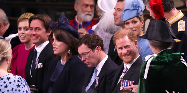 Prince Harry, seated, smiles in a suit as his aunt Anne, Princess Royal approaches him wearing a green jacket and dark hat with a red feather in it (with her back to the camera)