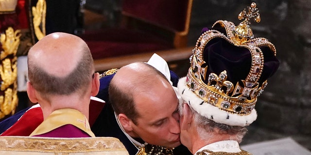 Prince William kisses his father's left cheek, who is sitting in a chair with a dark crown as he is crowned King