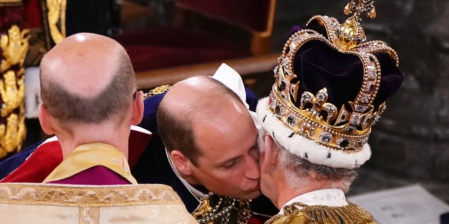 Prince William kisses his father's left cheek, who is sitting in a chair with a dark crown as he is crowned King