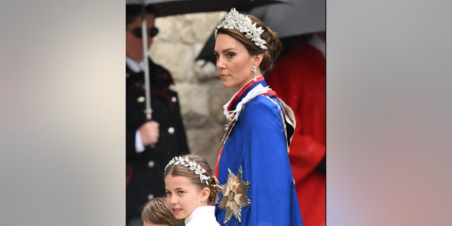 Kate Middleton in a leaf tiara walks in with a stoic expression to Westminster Abbey, wearing a blue cape, walking alongside her is Princess Charlotte in a matching, smaller tiara