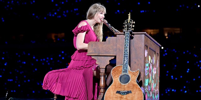 Taylor Swift in a pink dress plays behind a brown piano with a guitar leaning up against it at the Eras Tour