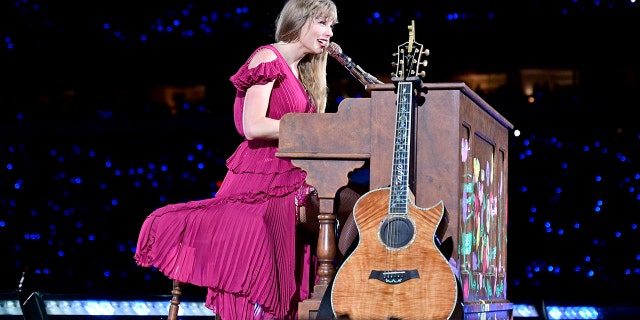 Taylor Swift in a pink dress plays behind a brown piano with a guitar leaning up against it at the Eras Tour