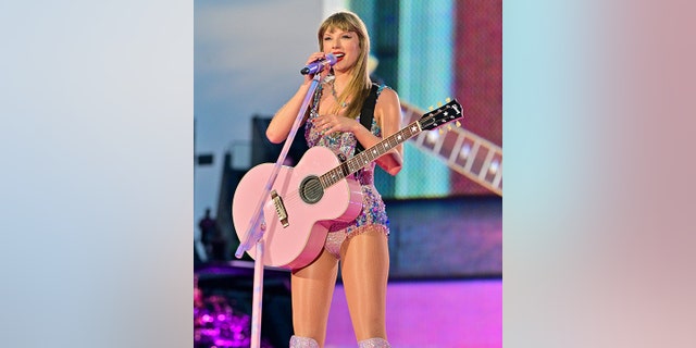 Taylor Swift holds the microphone during the Eras Tour playing a pink guitar