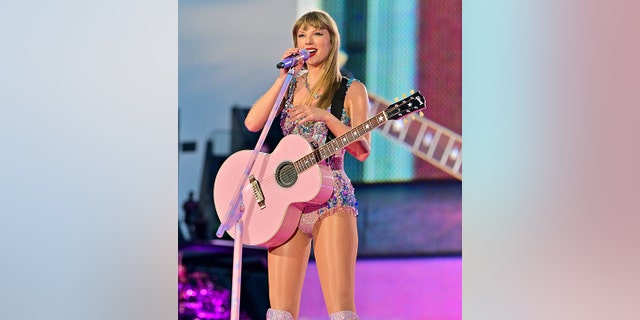 Taylor Swift holds the microphone during the Eras Tour playing on a pink guitar