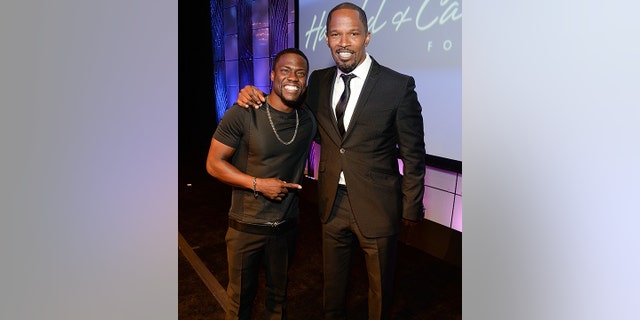 Kevin Hart wearing a black t-shirt and pants points to Jamie Foxx in a black suit and tie as they smile for a picture at an event