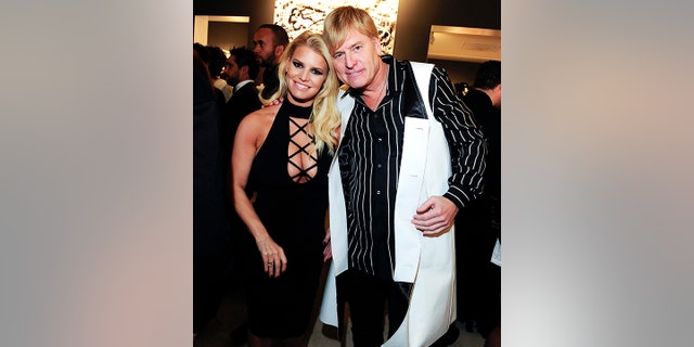 Jessica Simpson in a black dress with criss-cross cut outs at the top smiles next to her father Joe Simpson in a black striped shirt and white vest